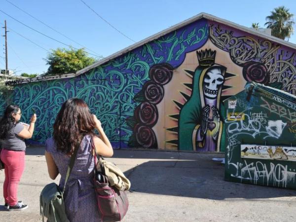Two people taking a photo of a mural on a building. The mural features a skeleton in a robe and crown with hands around a floating heart symbol.